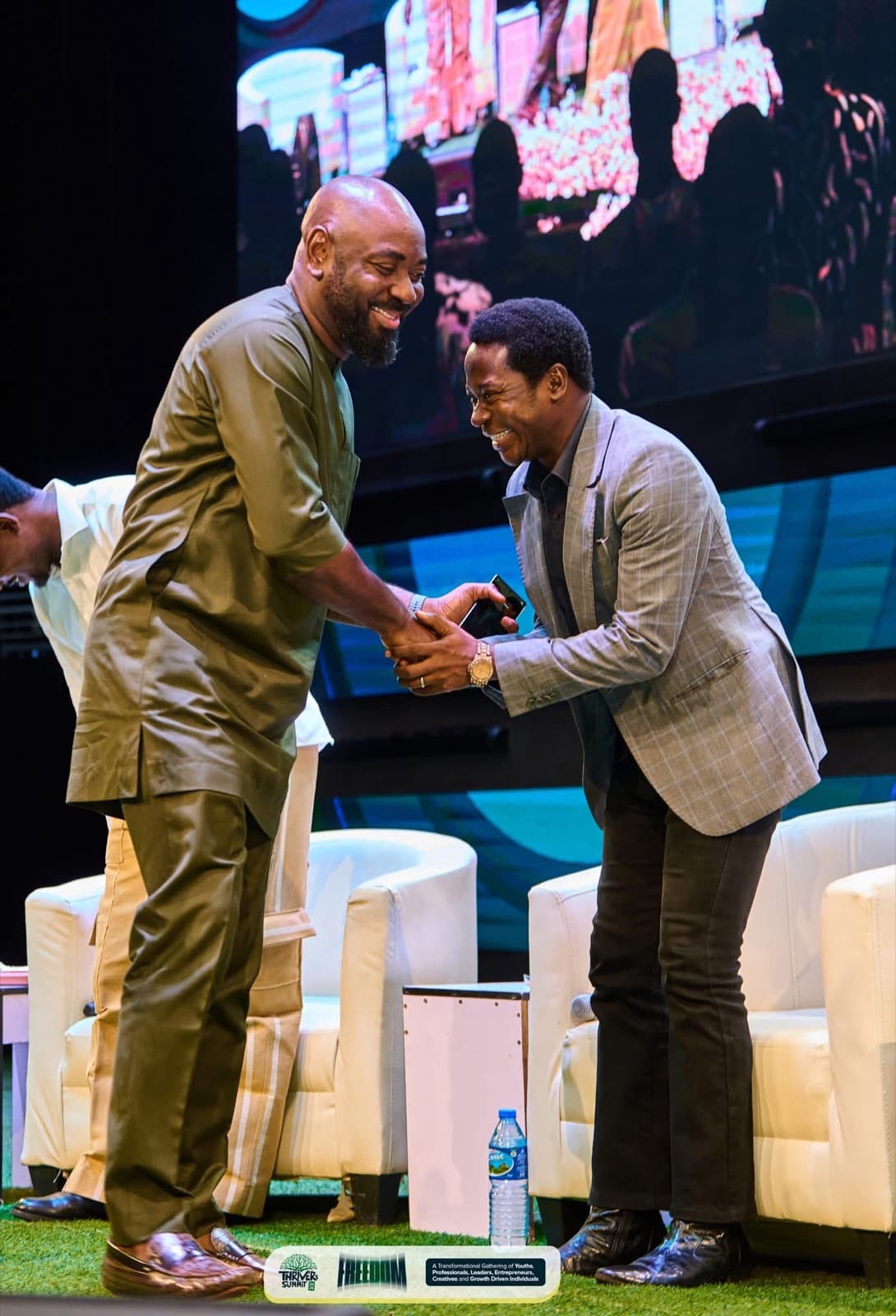 Two men shaking hands warmly on stage beside white armchairs