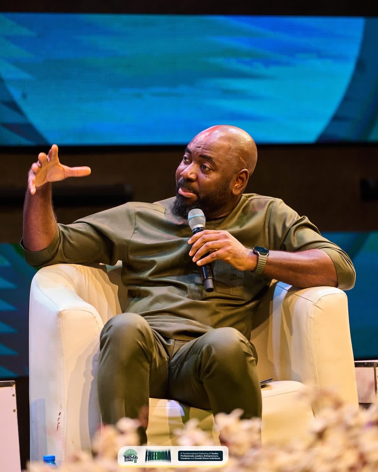 Speaker with a microphone seated in a white armchair, gesturing as he talks